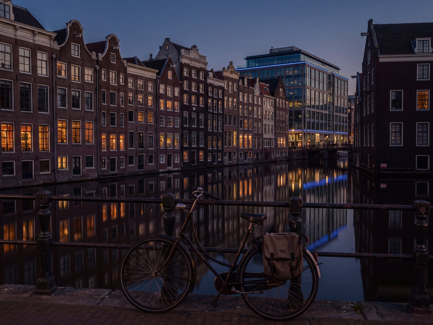 Amsterdam canal houses at blue hour, warm lit windows, a glass office building at the end of the row, a bicycle on the bridge